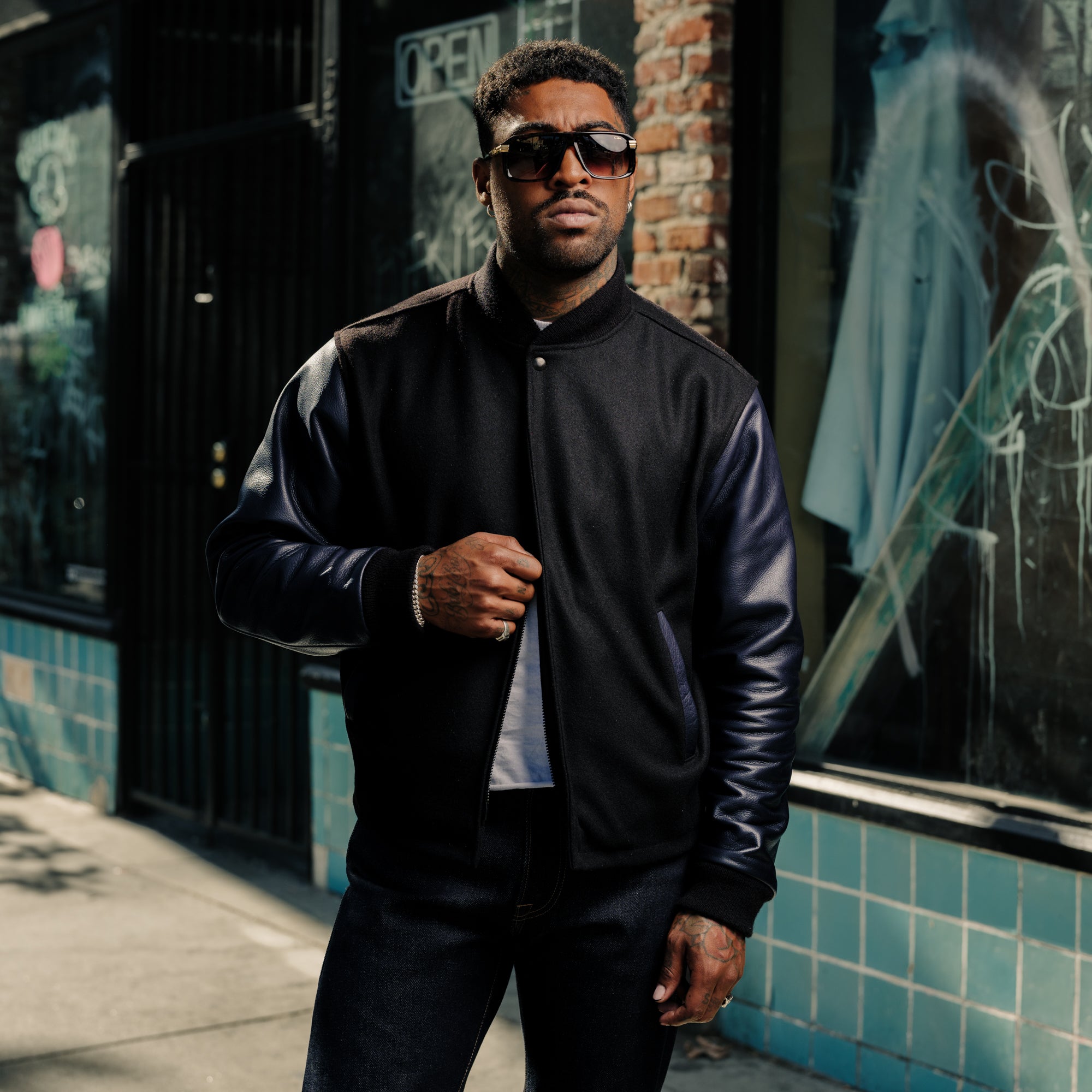 Man wearing a black jacket and sunglasses standing in front of a building with graffiti.