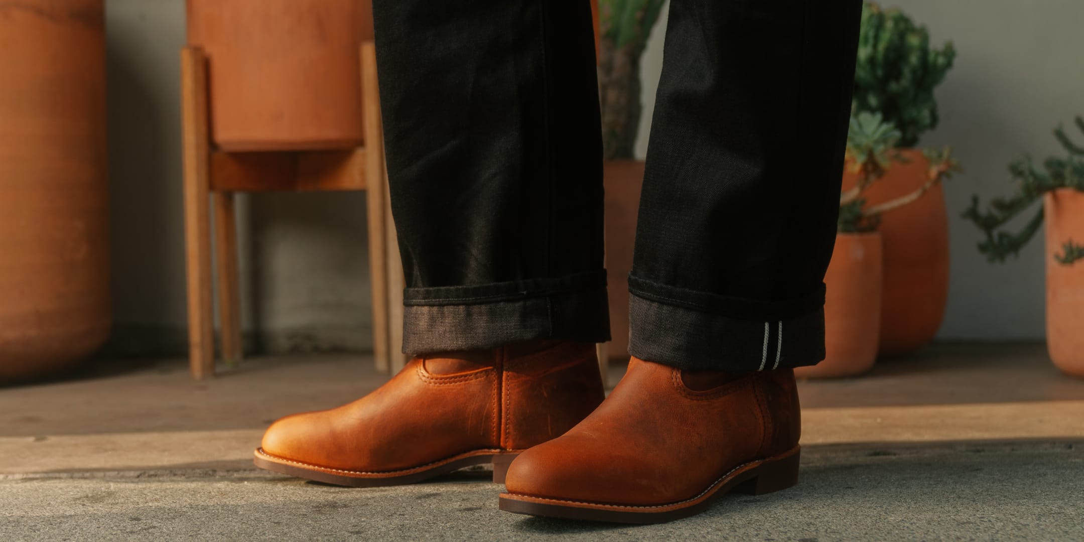 Person wearing brown leather boots and dark jeans standing indoors with plants in the background