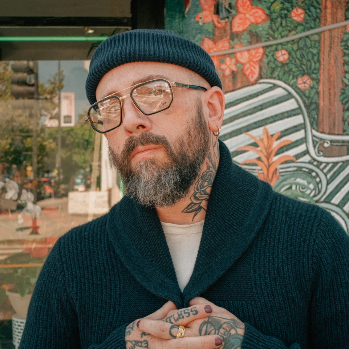 Man with tattoos wearing glasses and a dark sweater in front of a colorful mural.