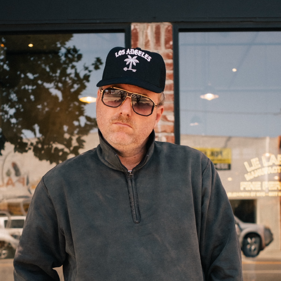 Man wearing a dark sweatshirt, cap and sunglasses standing in front of a store window.