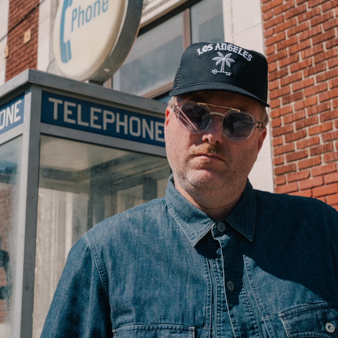 Man wearing a denim jacket and sunglasses in front of a vintage telephone booth.