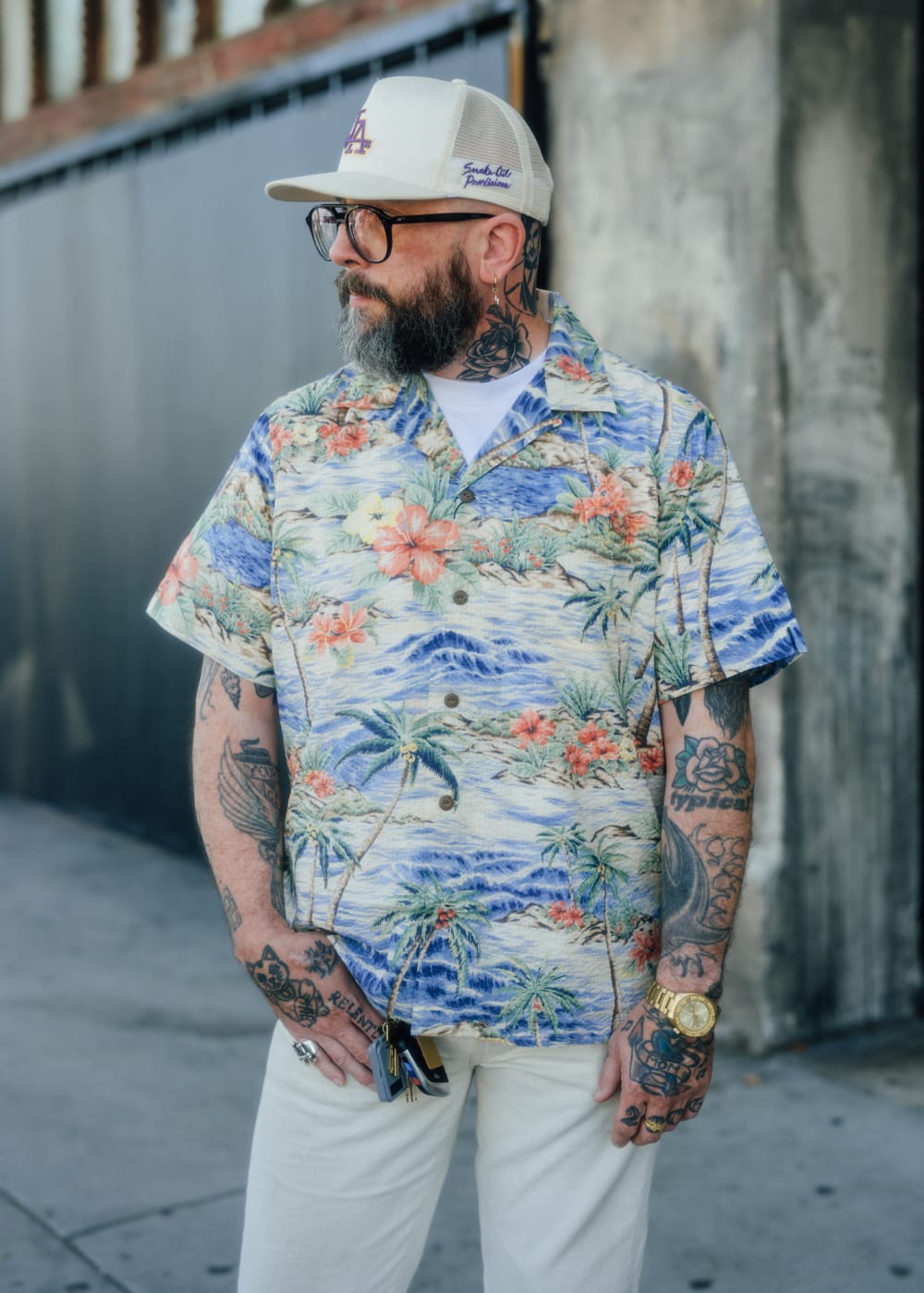 A man with a beard and tattoos wearing a colorful Hawaiian shirt with a blue ocean and tropical floral print, paired with a beige cap and white pants, standing outdoors against a concrete wall.
