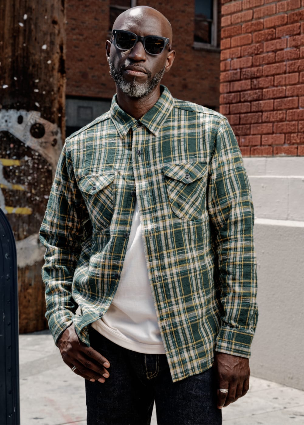 A man wearing a green and yellow plaid button-down shirt over a white t-shirt and dark jeans, standing outdoors in front of a brick wall.