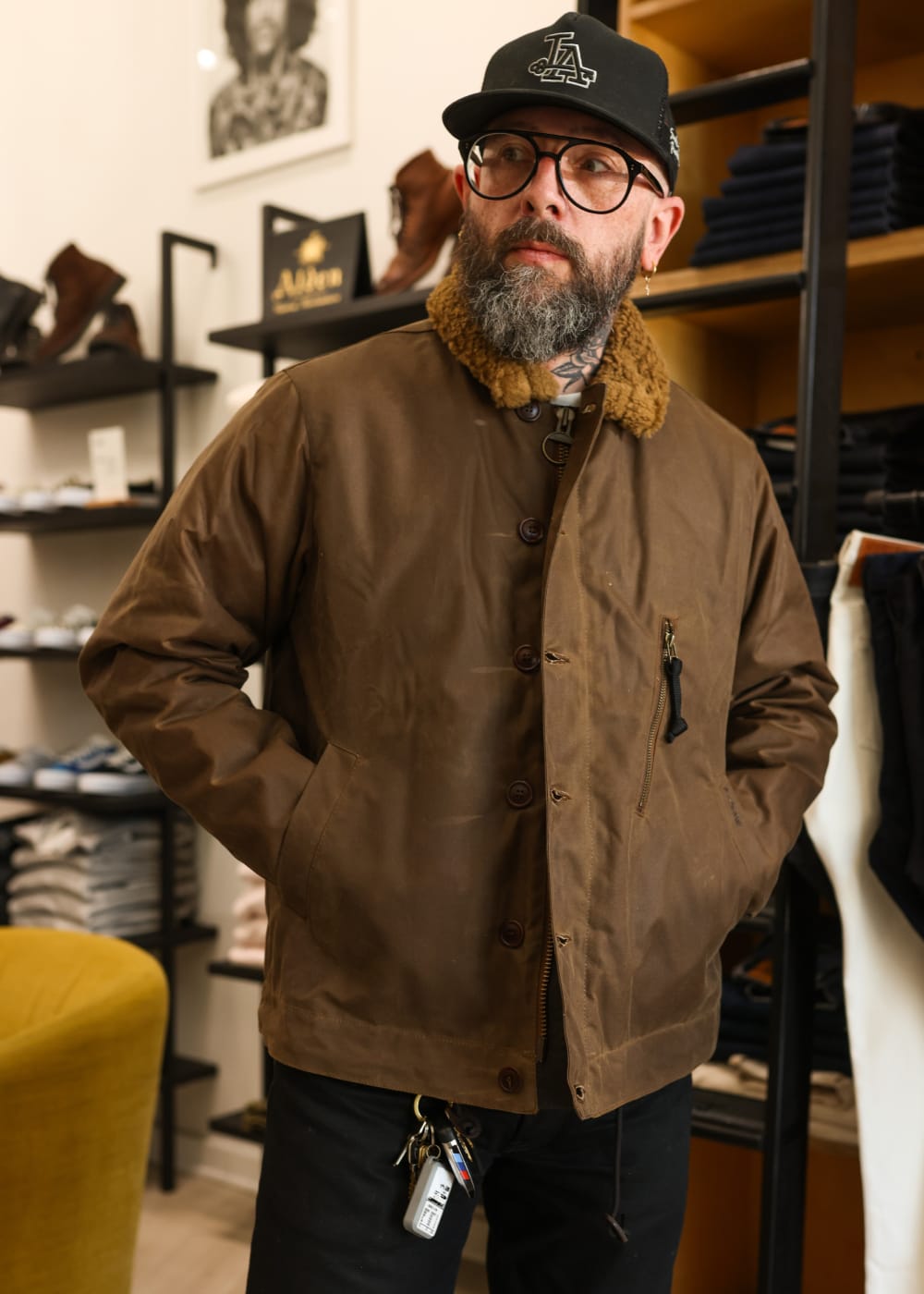 A man wearing a brown waxed cotton utility jacket with a tan shearling collar, black pants, and a black baseball cap, standing inside a clothing store.