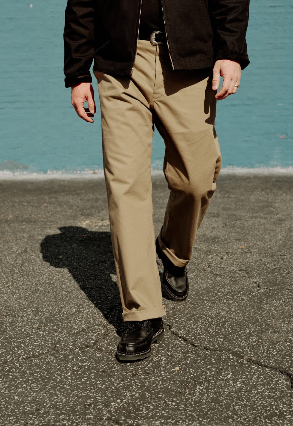 A person walking outdoors wearing tan chinos, a black jacket, and black shoes, with a blue water wall and gray pavement in the background.