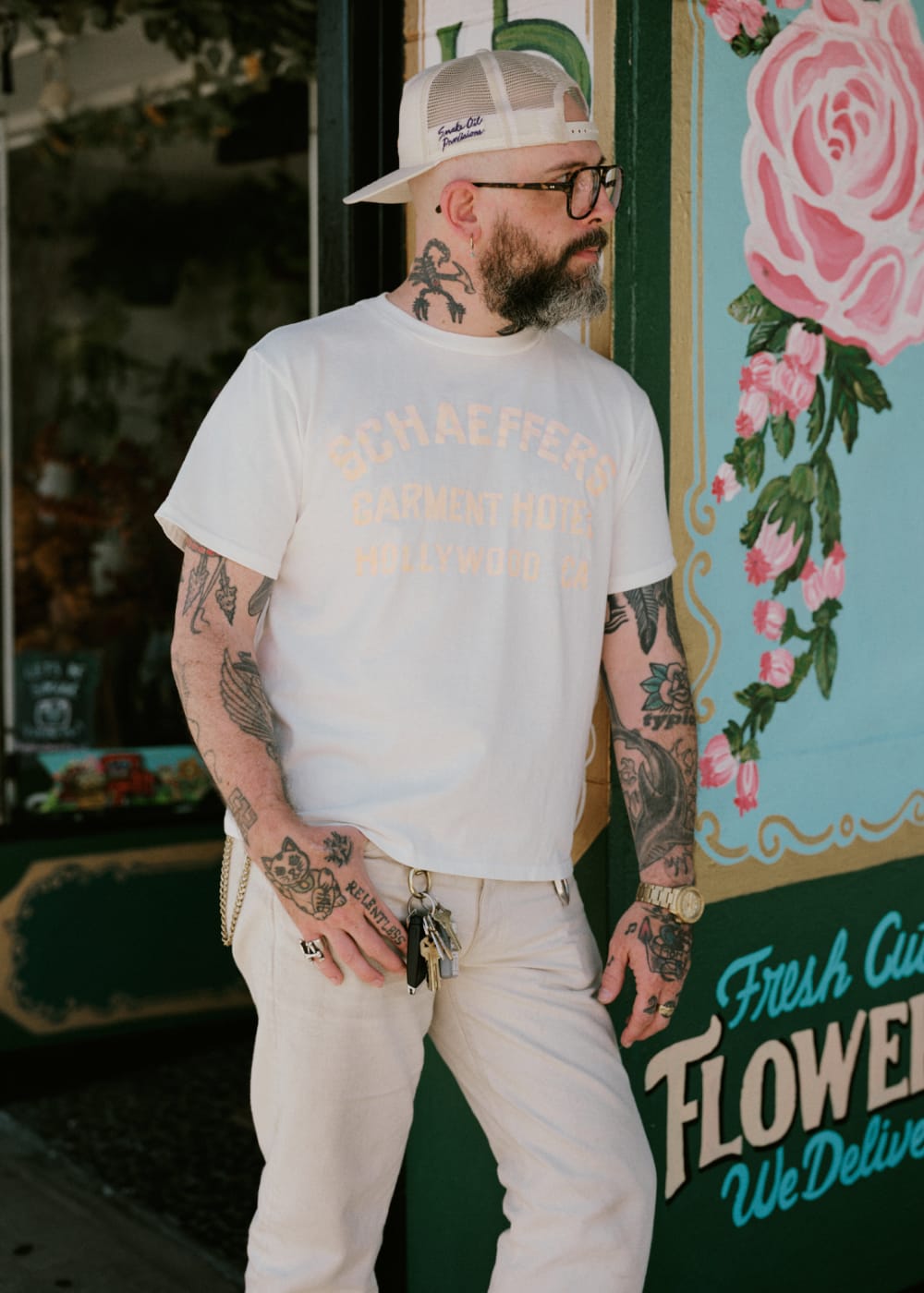 A man wearing a white graphic t-shirt with pale yellow text, cream pants, and a white cap, standing in front of a vintage-style flower shop sign.