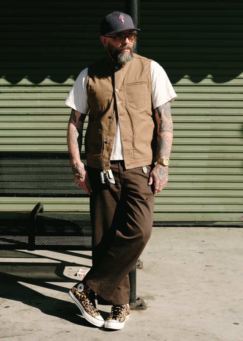 a bearded man with tattoos wears a brown vest over a white t-shirt and a black cap against a dark green shutter
