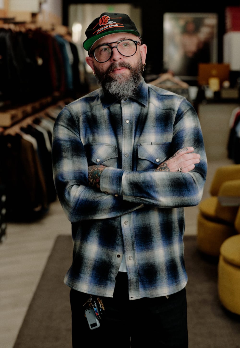 A man with a beard, glasses, and a cap stands with arms crossed inside a clothing store, wearing a blue and gray plaid button-down shirt.