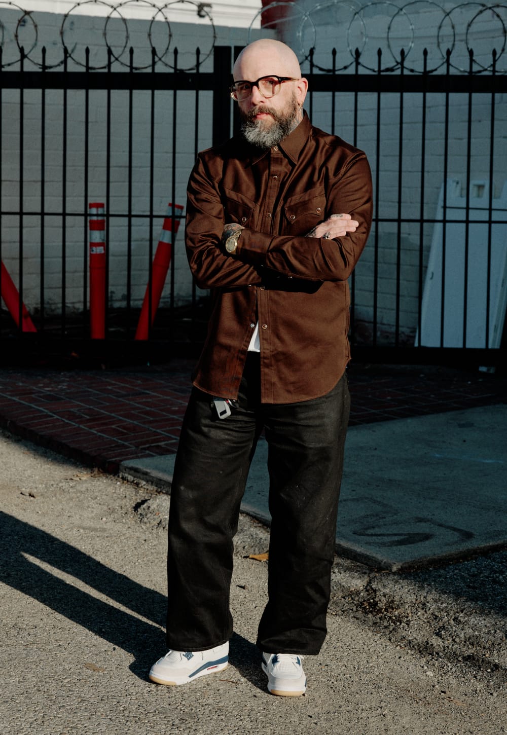 A man with a bald head, glasses, and a beard stands outdoors with his arms crossed, wearing a brown button-up shirt, dark jeans, and white sneakers. He is standing in front of a black metal fence with barbed wire on a concrete ground.