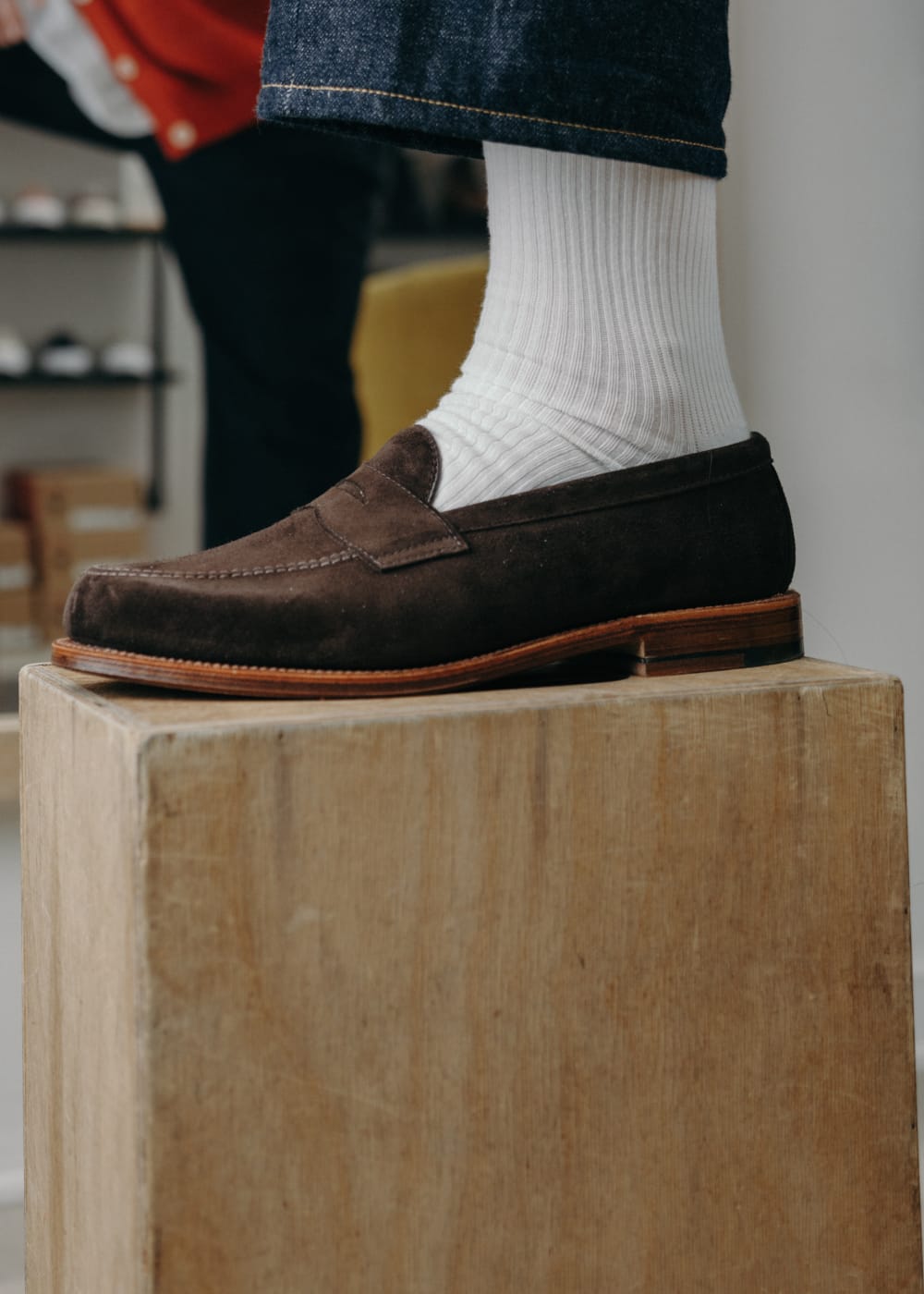 Brown loafer worn with white socks on a wooden block, with blurred background