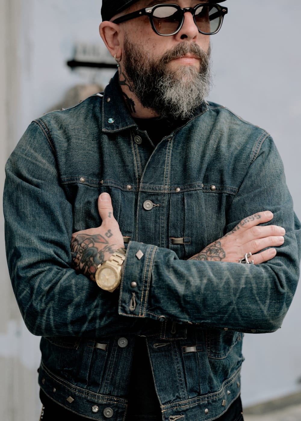 A man with a gray beard, tattoos, and sunglasses wearing a dark blue distressed denim jacket, standing with his arms crossed.