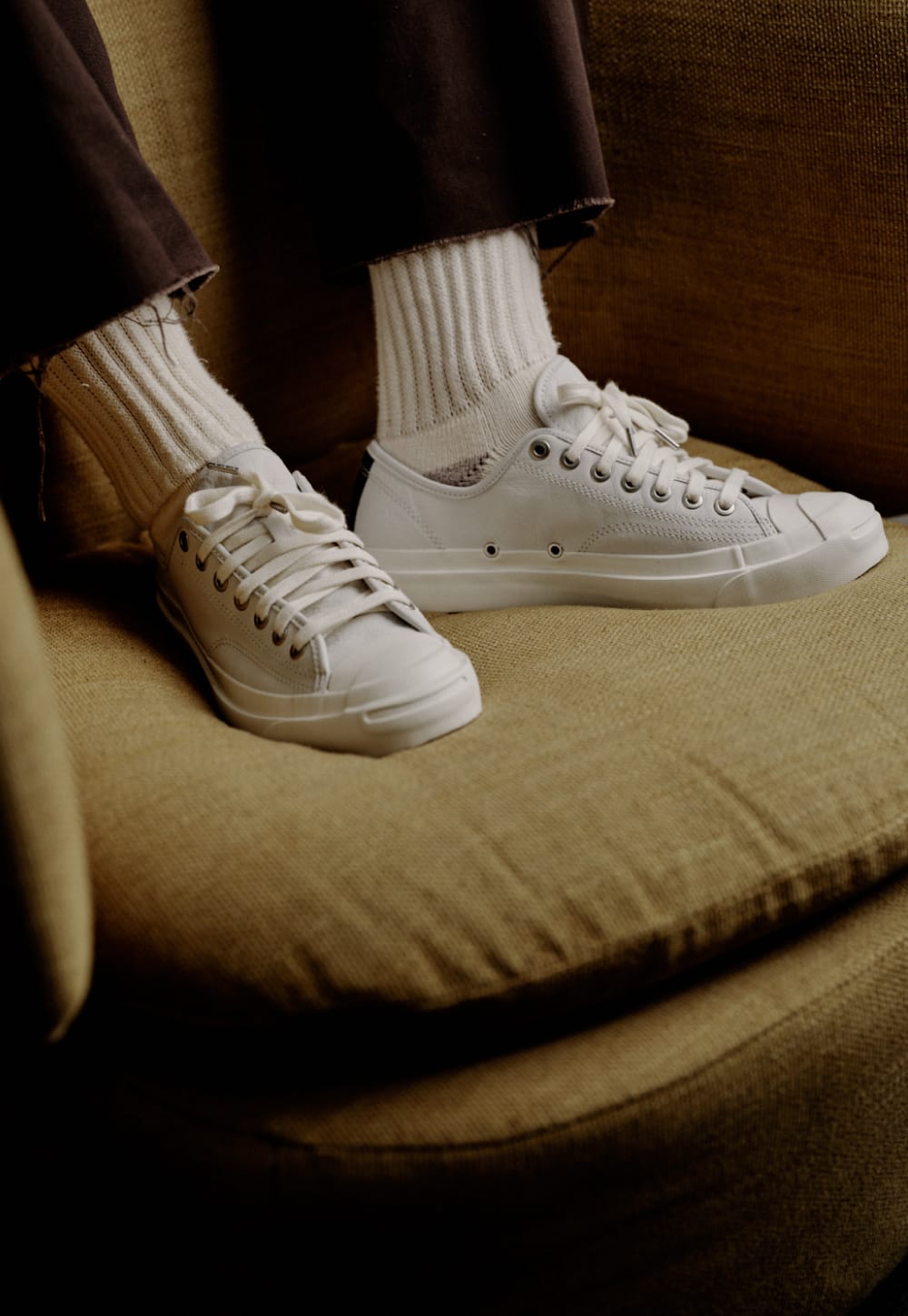 White low-top canvas sneakers with white ribbed crew socks, worn by a person sitting on a textured olive-brown upholstered chair.