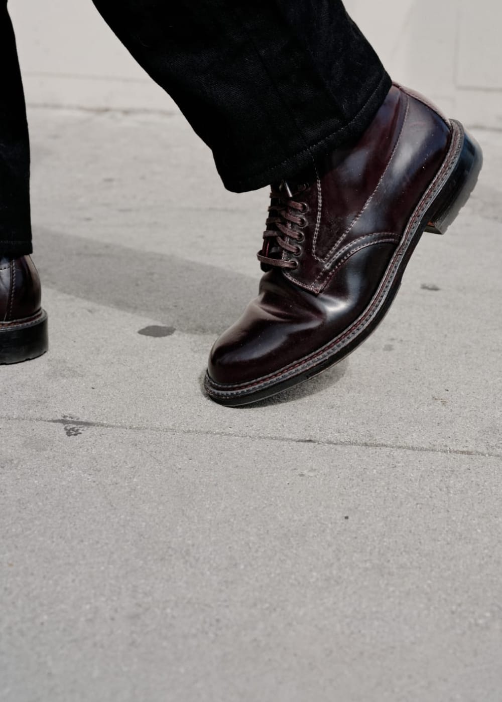 A close-up of a person's legs wearing dark brown leather lace-up boots and black pants, standing on a gray concrete pavement.