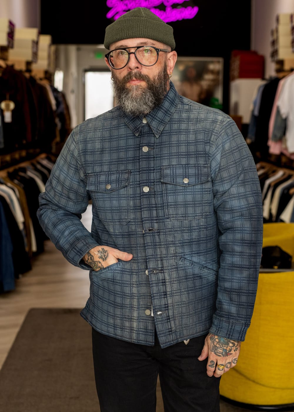 A man with a beard, glasses, and tattoos wearing a blue and gray plaid button-up shirt, black pants, and an olive green beanie, standing inside a clothing store.