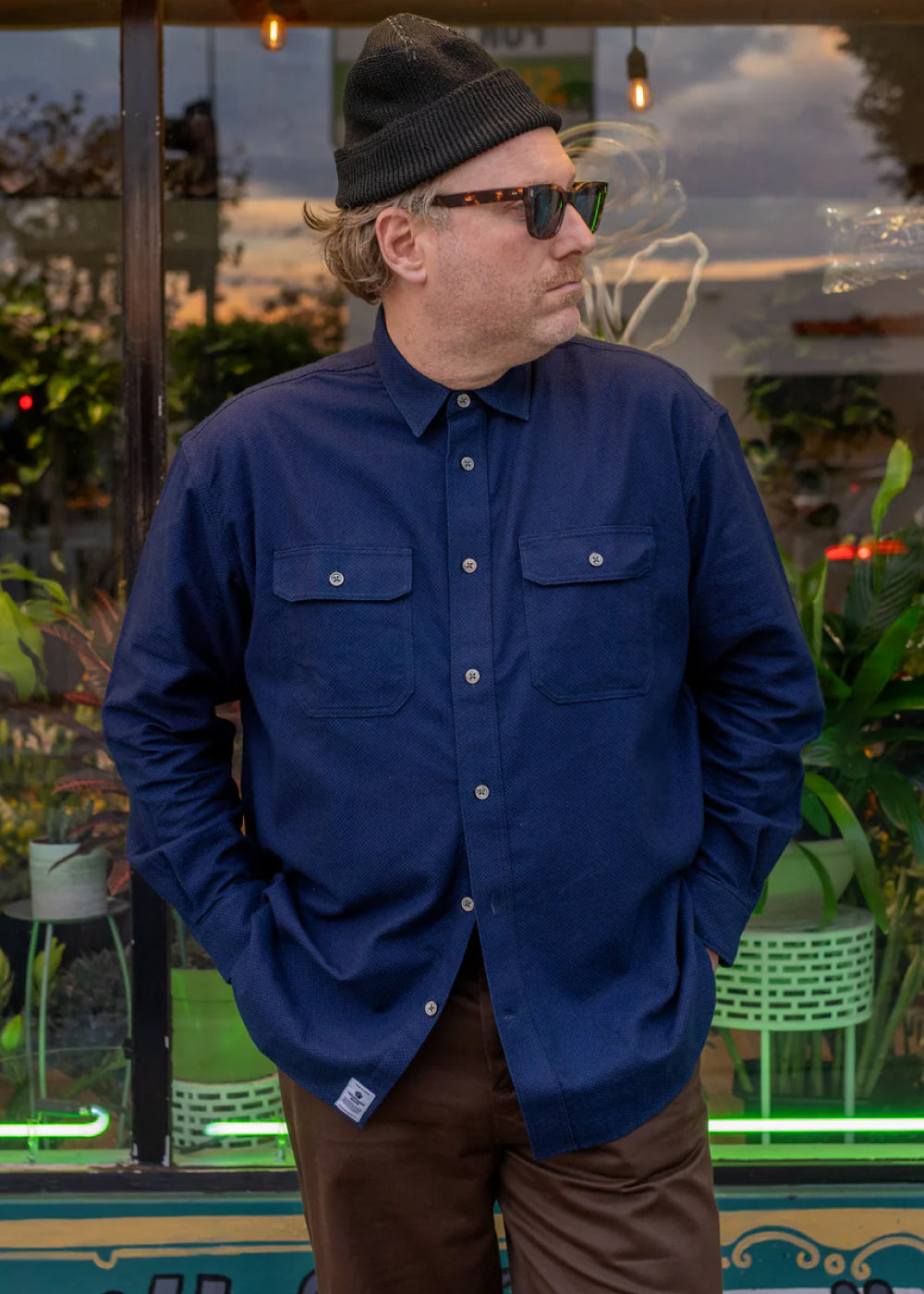 Man wearing a blue shirt, black beanie, and sunglasses standing in an outdoor setting with plants and neon lights.