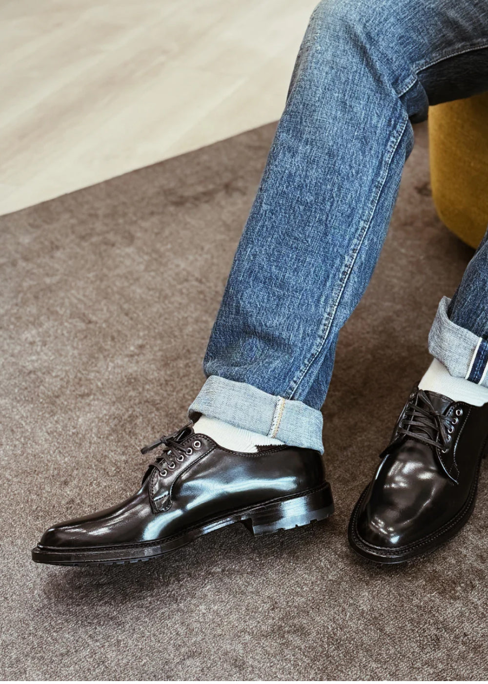Black dress shoes worn with rolled-up blue jeans on a carpeted floor.