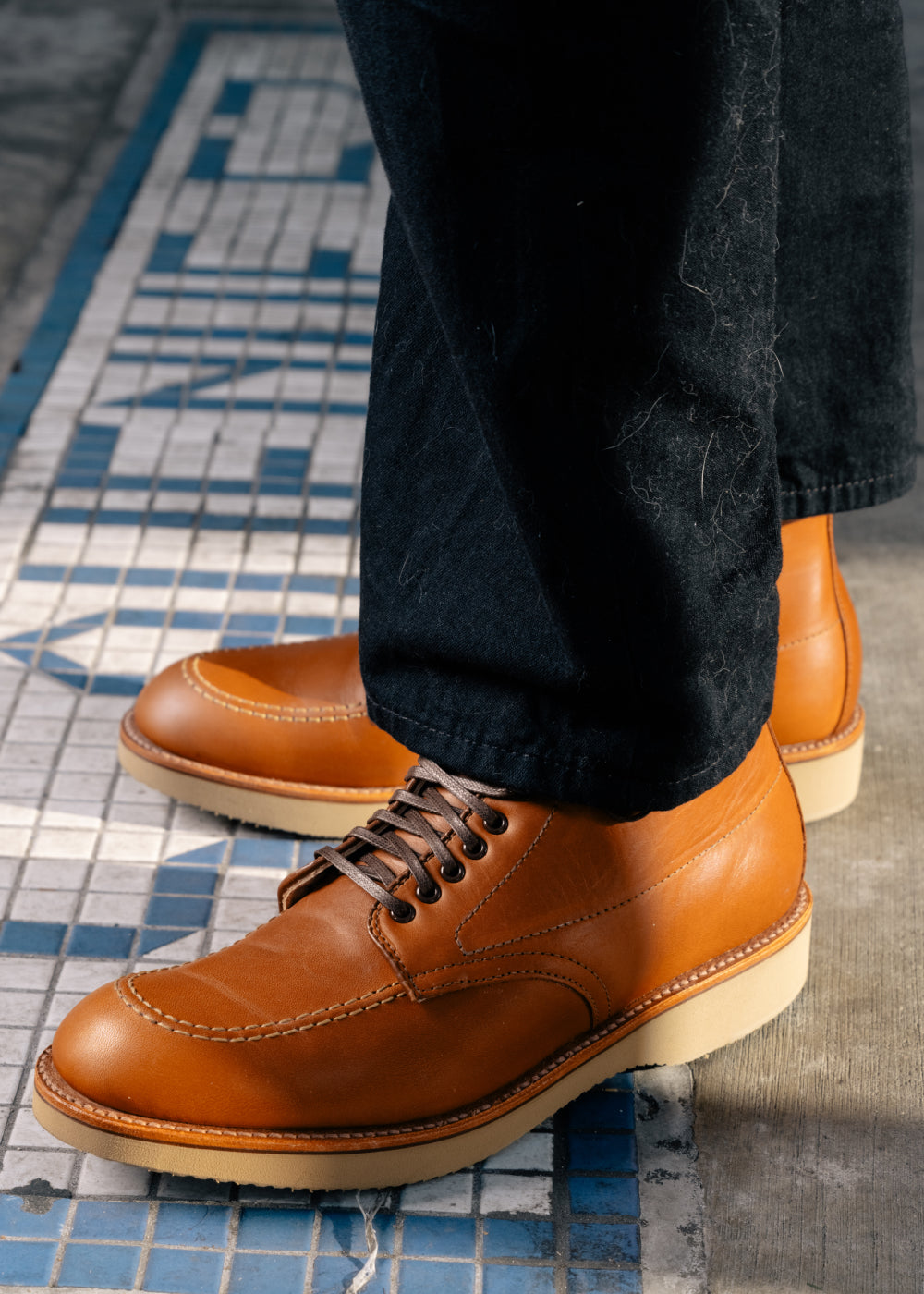 Brown leather shoes worn with dark jeans on a patterned floor.
