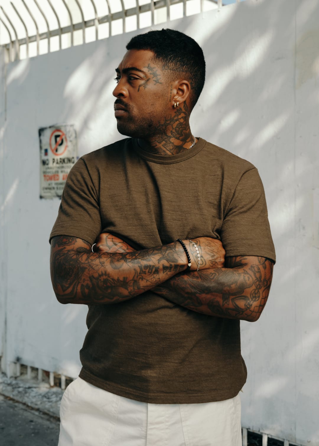 Man with tattoos wearing a brown t-shirt and white pants standing against a white wall.