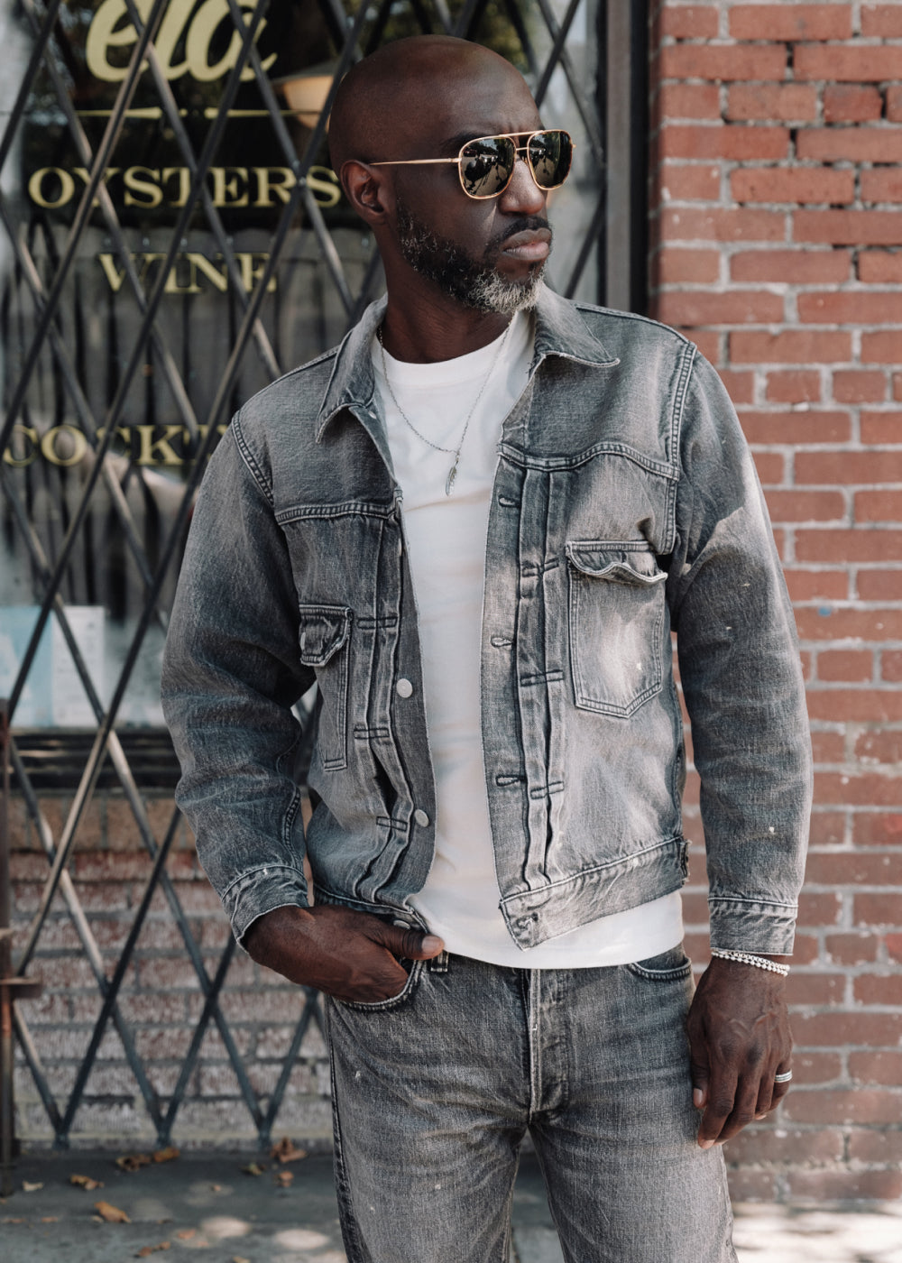 Man wearing a denim jacket and sunglasses standing in front of a brick wall.