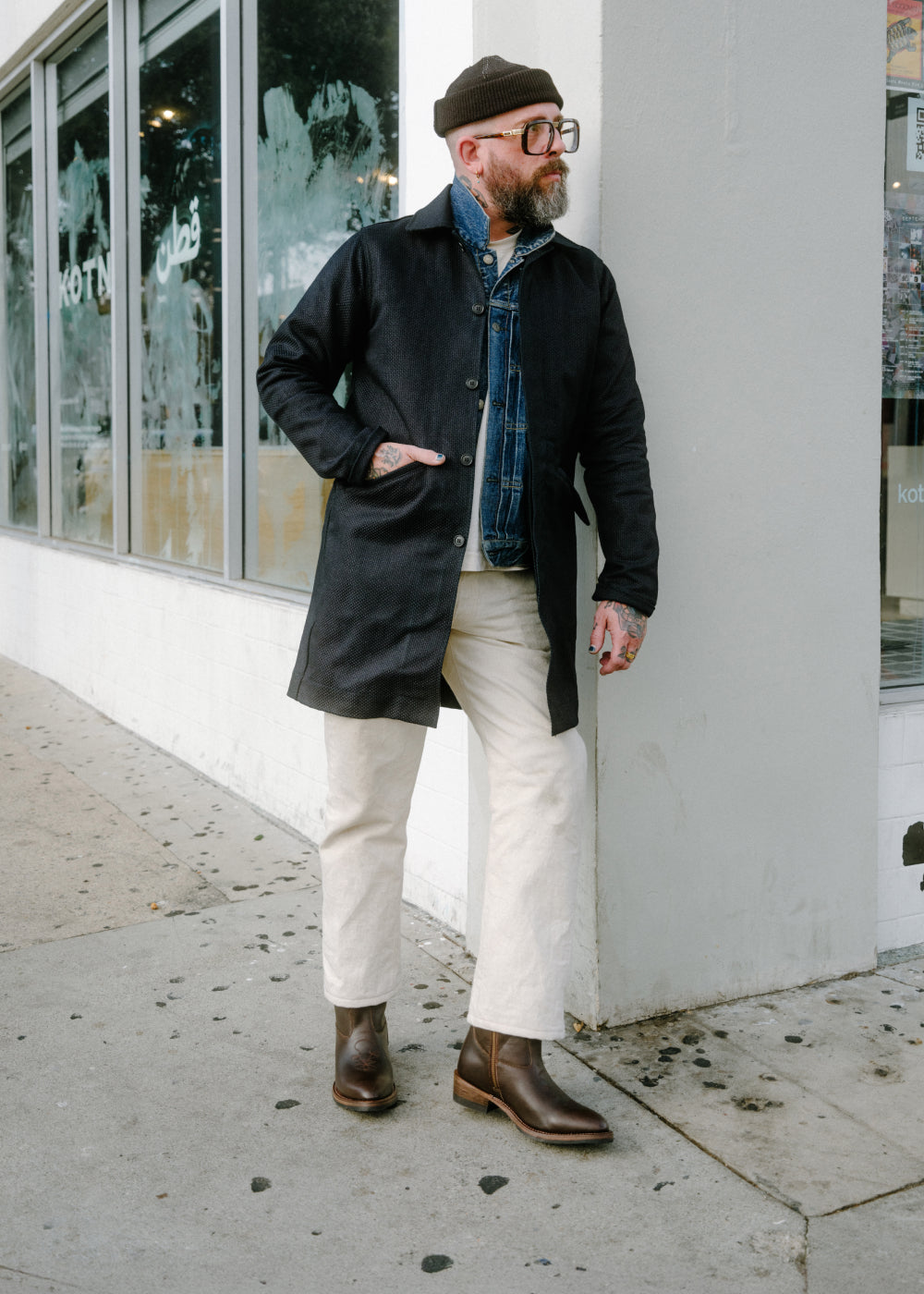 Man wearing a black coat, white pants, and brown boots standing on a sidewalk.
