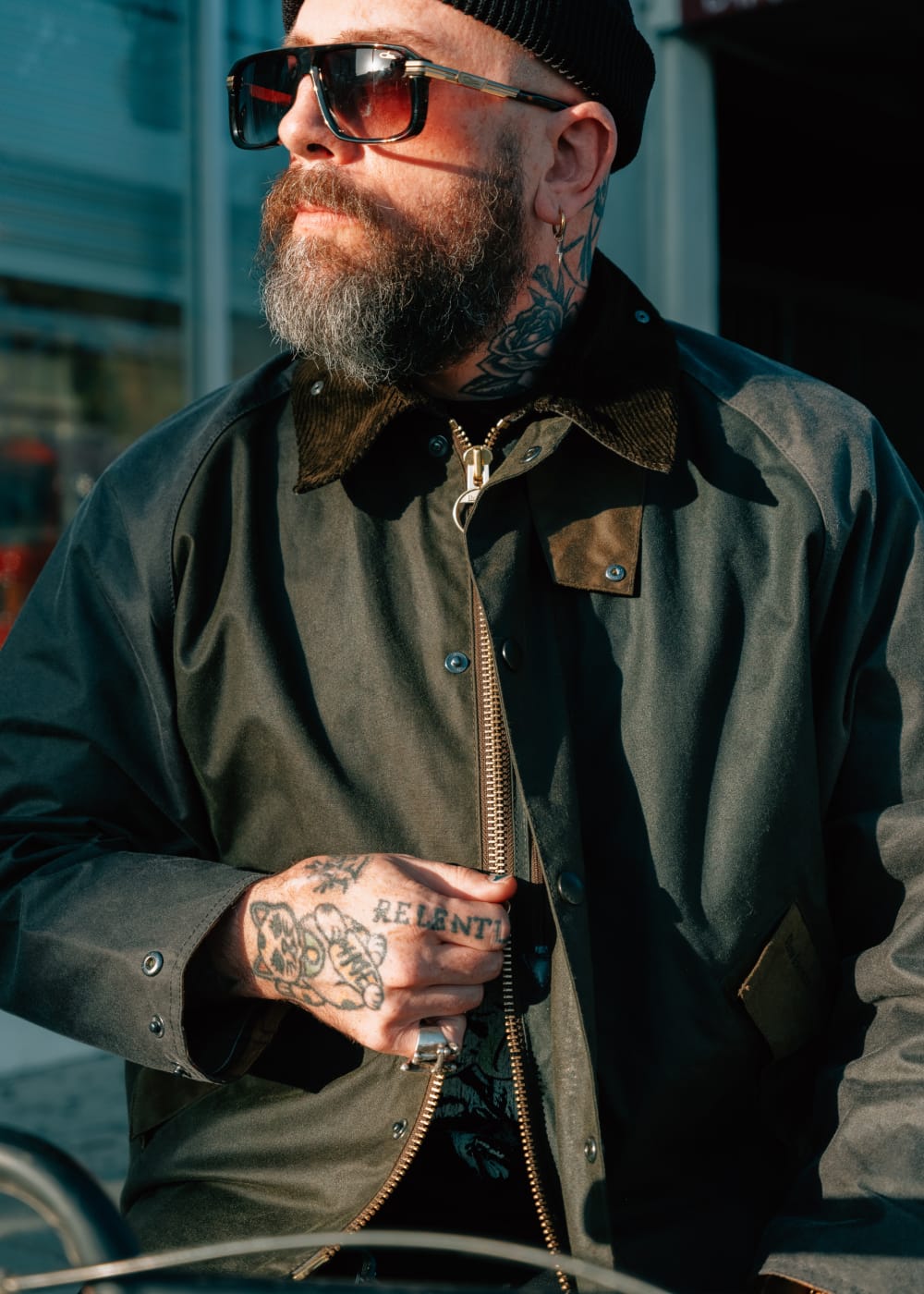 Man wearing sunglasses and a dark coat with tattoos on his arm, standing outdoors.