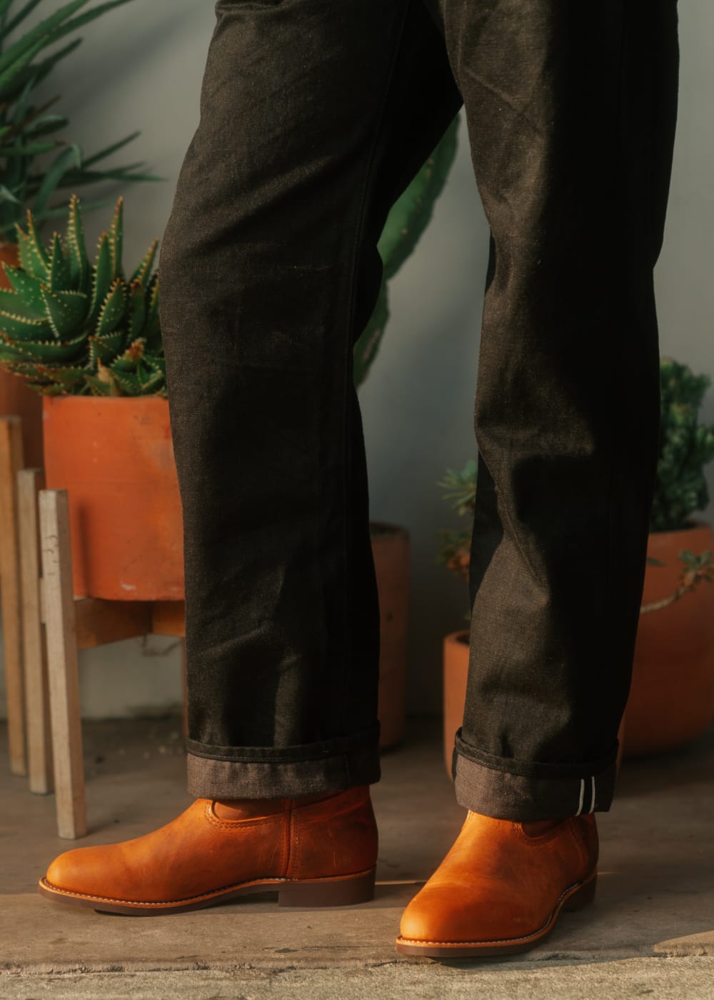 Person wearing brown leather boots and dark jeans standing indoors with plants in the background