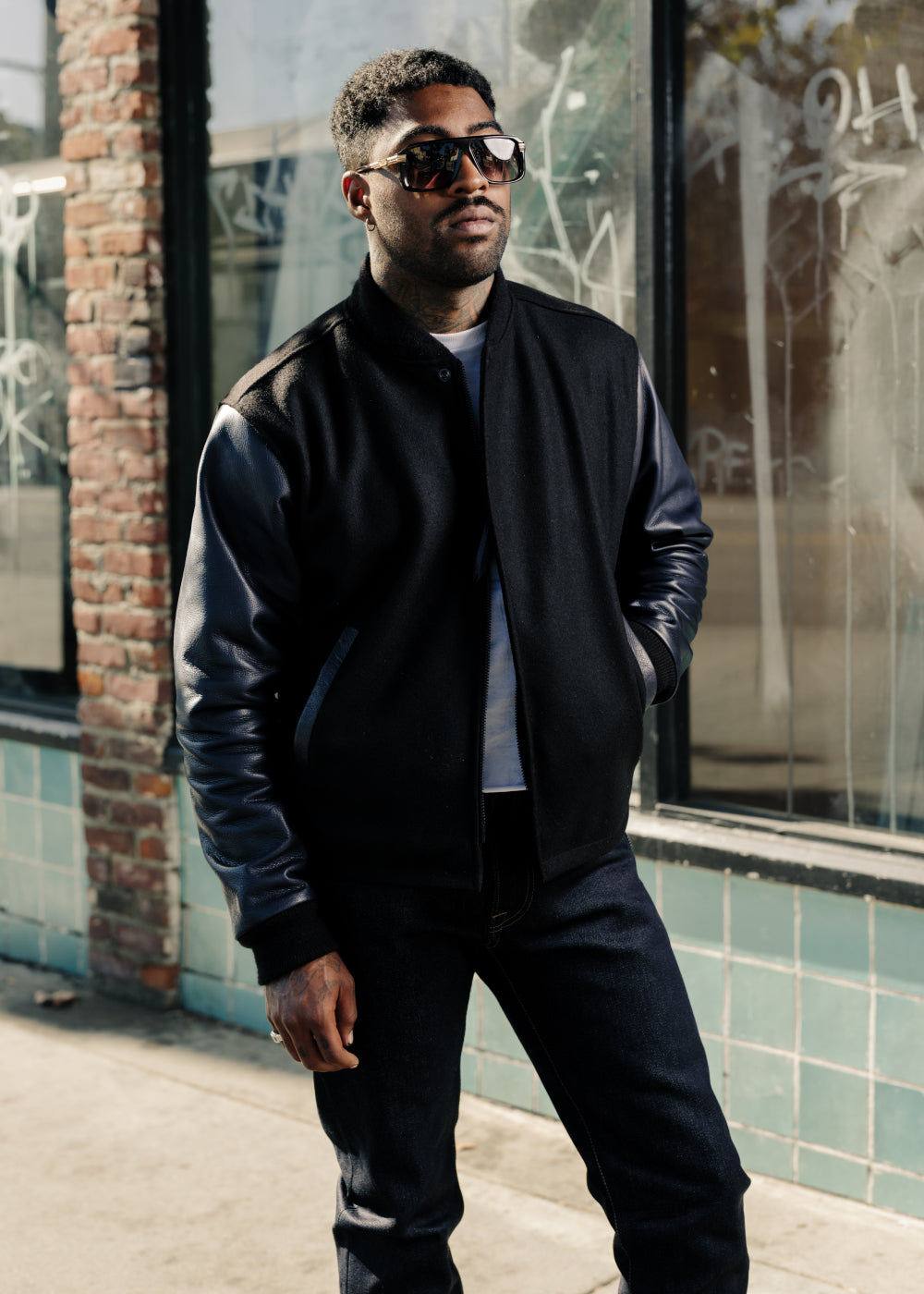Man wearing a black bomber jacket and sunglasses standing in front of a building with glass windows.