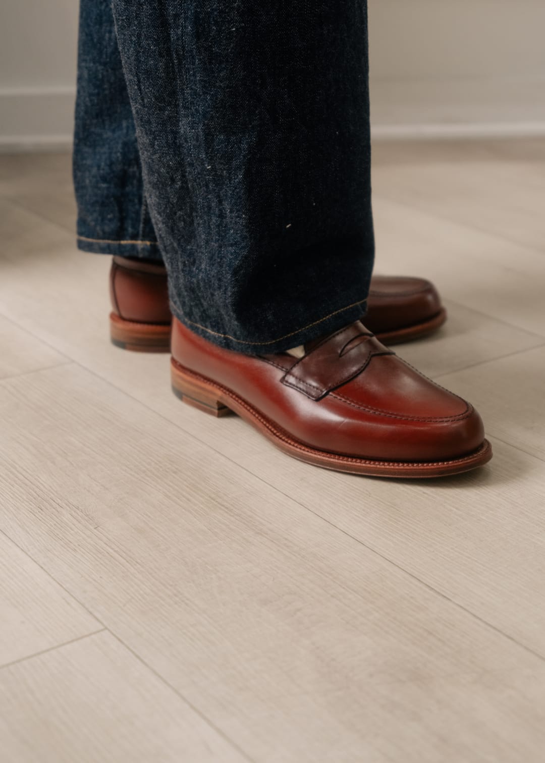 Brown loafers worn with dark jeans on a light wooden floor.