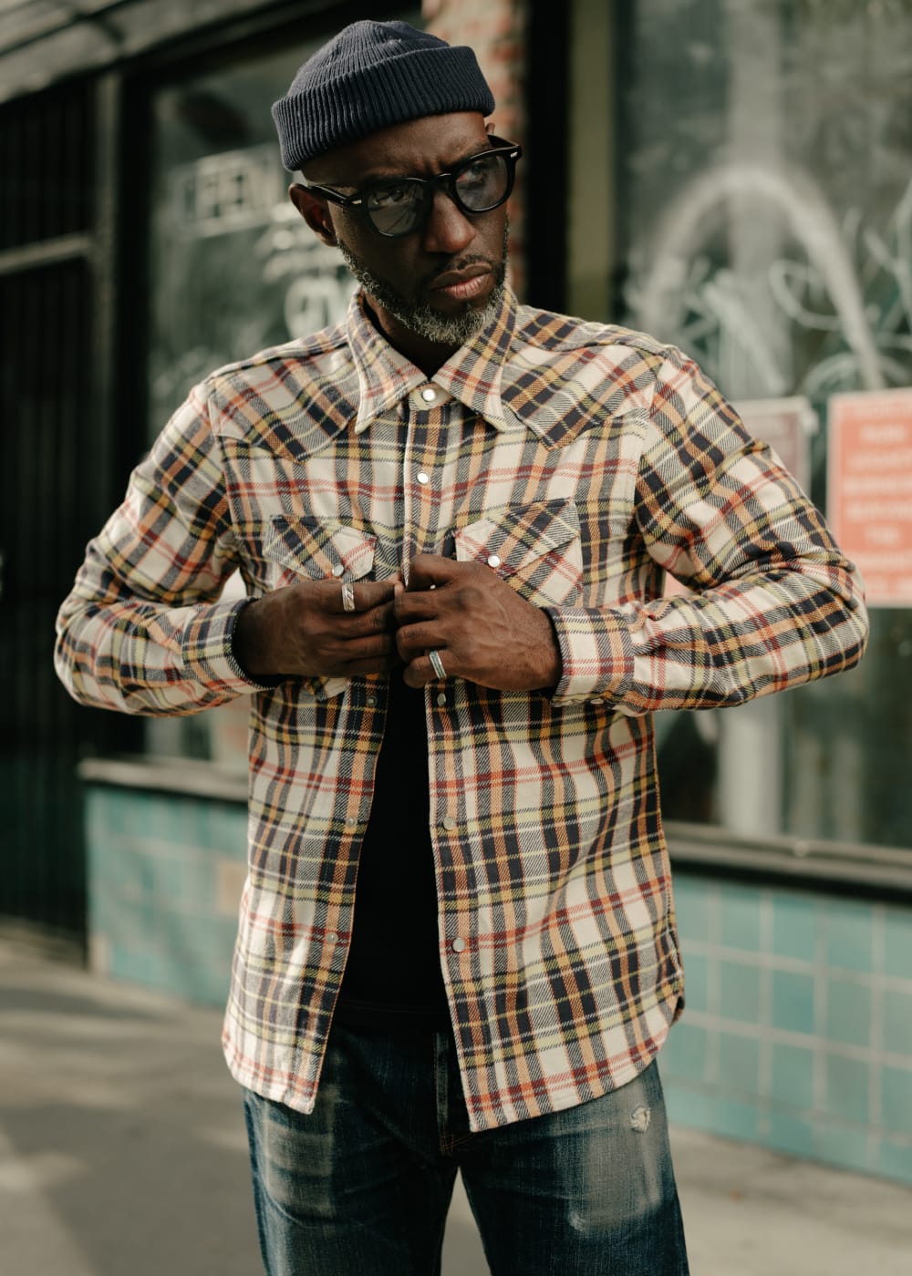 Man wearing a plaid shirt and beanie standing in front of a building with graffiti.