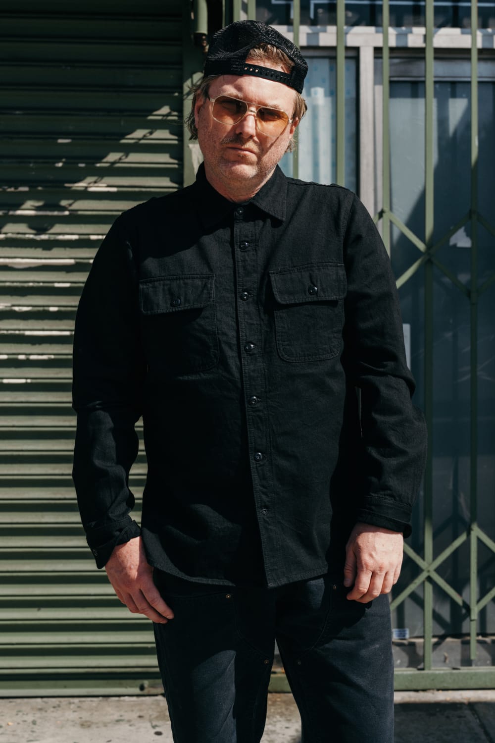 Person wearing a black shirt and sunglasses standing in front of a green metal gate.