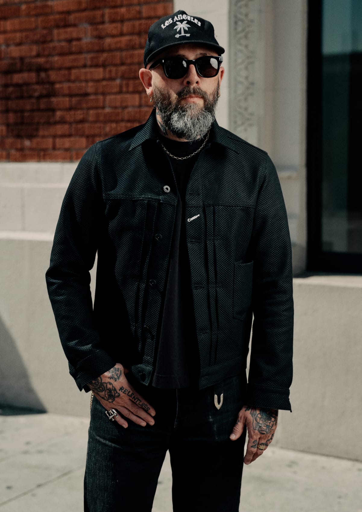 A man with a beard and tattoos wearing a black textured button-up jacket, black t-shirt, black jeans, a black baseball cap with a white graphic, and sunglasses, standing outdoors against a brick wall and concrete building.