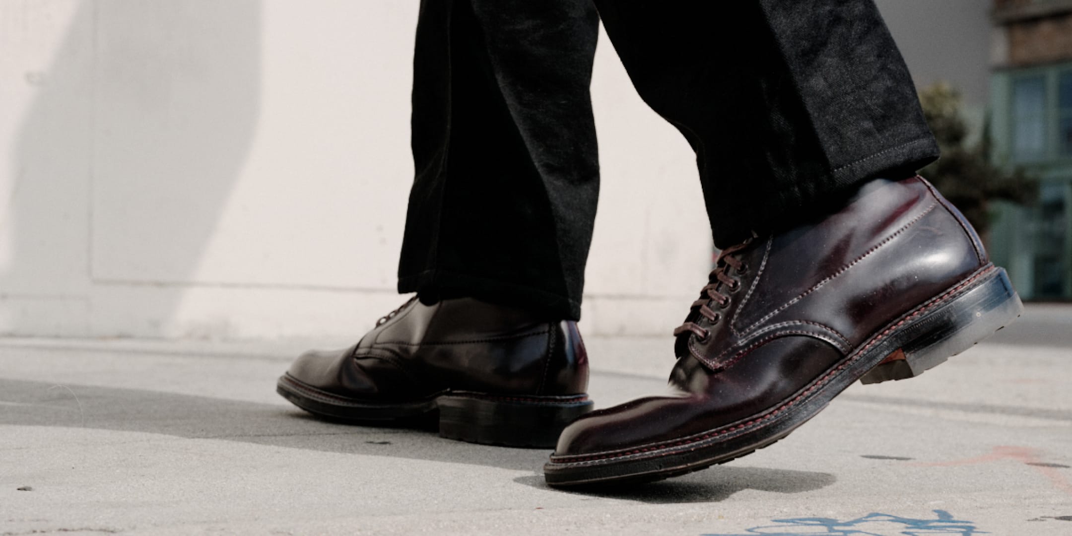 A close-up of a person's legs wearing dark brown leather lace-up dress shoes with black trousers, standing on a concrete ground.